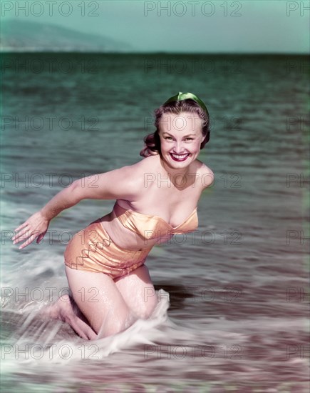 1940s SMILING BLOND WOMAN WEARING GOLD TWO PIECE BIKINI BATHING SUIT KNEELING IN OCEAN SURF LOOKING AT CAMERA