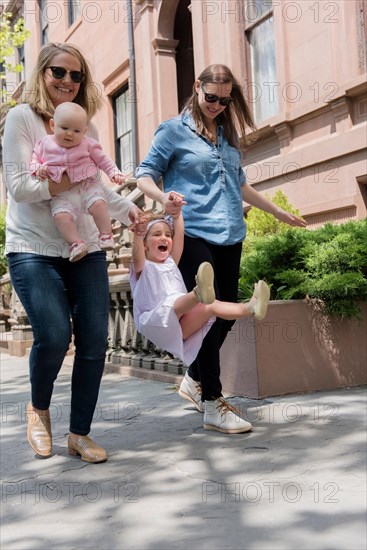 Caucasian mothers swinging daughter on city sidewalk