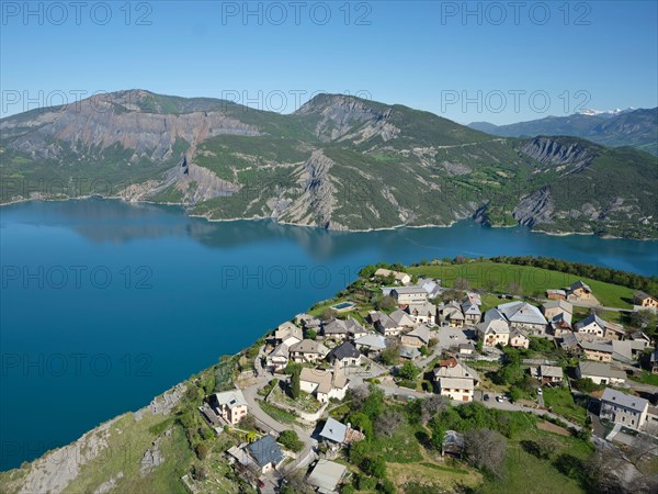 AERIAL VIEW - Village perched 250m-high above Lake Serre-Poncon. Le Sauze-du-Lac, Hautes Alpes, France.