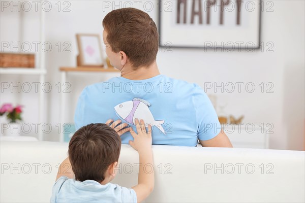 Little boy sticking paper fish to his father's back indoors. April fool's day prank