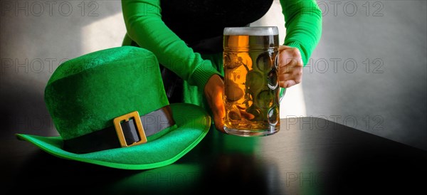 Woman's hand holding glass of beer near St. Patrick's day hat of a leprechaun on the black background