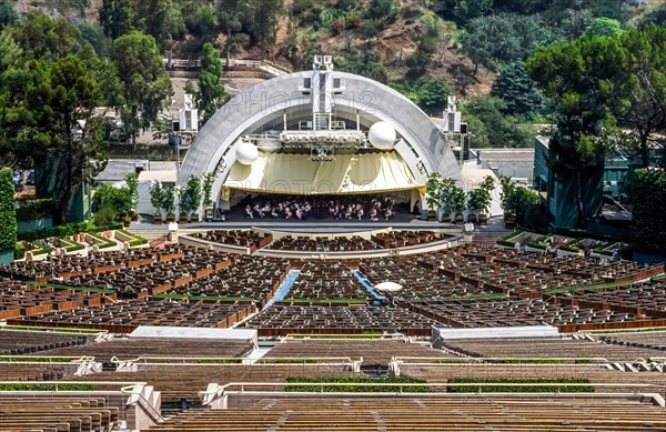 An orchestra rehearses on stage before 17,376 sloping empty seats in the famed Hollywood Bowl that is named for a natural amphitheater created by the Hollywood Hills in Hollywood, California, USA. An awning protects the musicians from the sun during their daytime practice. This semicircular shell above the stage dates to 1929, and shows large white spheres that were added later to improve acoustics. A new, slightly larger shell was installed in 2004. This outdoor showplace is home to the Hollywood Bowl Orchestra and Los Angeles Philharmonic and hosts other musical events throughout the year.