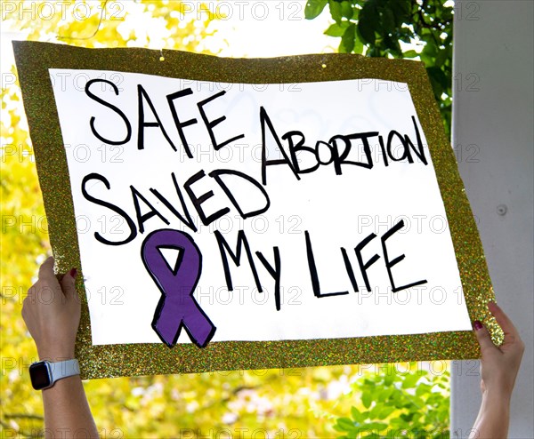 Sign with glitter edge reads Safe Abortion Saved My Live - blurred outdoor background at Reproductive Justice Rally