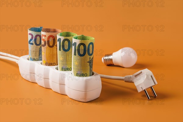 Rising energy costs concept. Euro money banknotes plugged in a white power strip, electrical plug and light bulb over orange background. Energy costs.