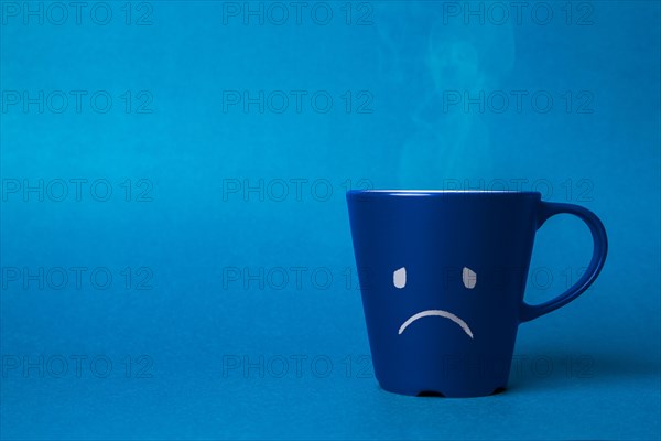 Stock photo of a blue cup on a blue background with a sad face drawn. Blue monday concept
