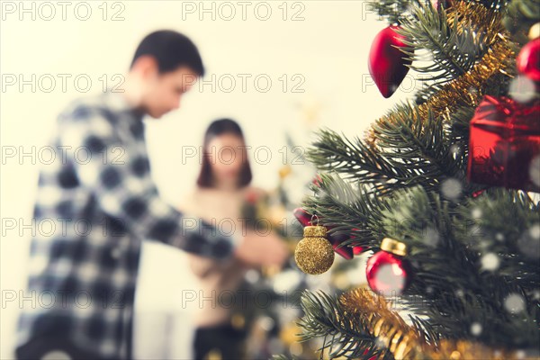 Close up of ornaments on Christmas tree with blur couple decorating another tree in background