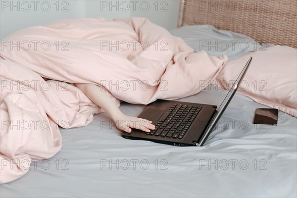 Bored funny child lying in bed under blanket and learning in virtual online school class. Hands typing on computer keyboard. Kid working on laptop