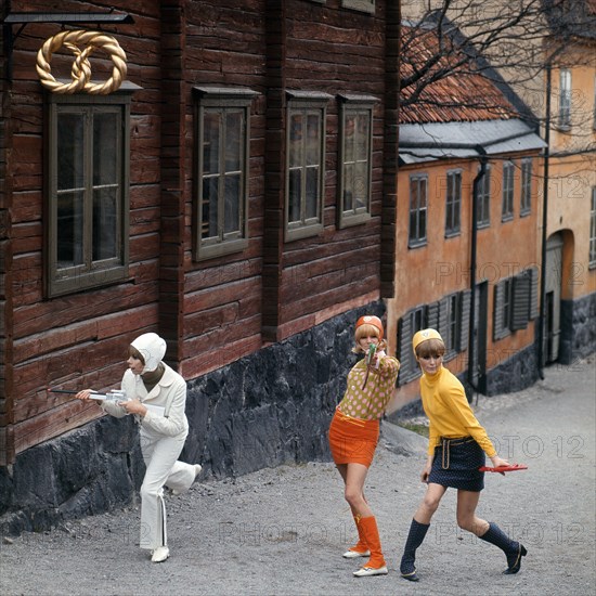 Three young women in brightly coloured 1960's style dresses  attending to do a hold up carrying plastic guns. photo: Bo Arrhed