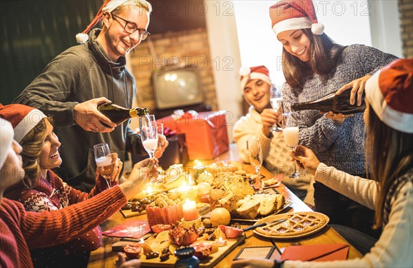 Friends group with santa hats celebrating Christmas with champagne and sweets food at home dinner - Winter holidays concept with people enjoying time