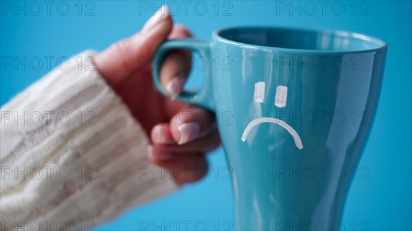 Blue Monday concept. Close-up of a female hand in a white sweater holds a blue cup with a sad smiley as a symbol of the most depressive day of the yea