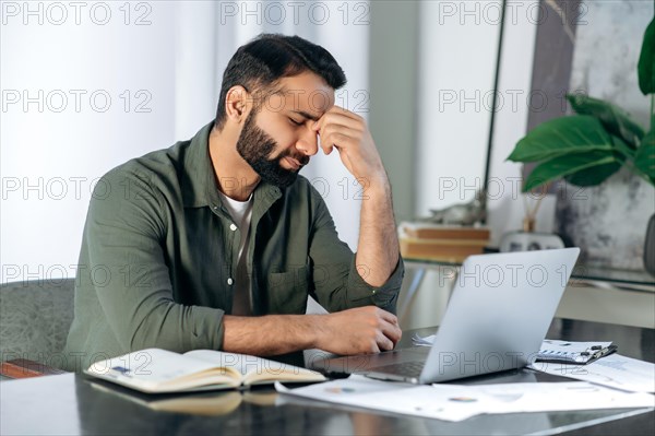 Tired exhausted arabic or indian man, office worker, manager or freelancer, sitting at his desk, tired of working in a laptop, overworked, having a headache, closed his eyes, needs rest and break