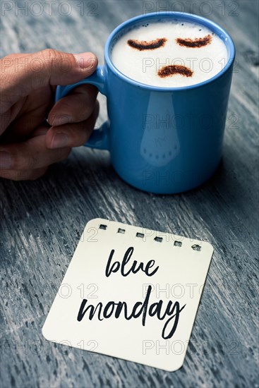 closeup of a note with the text blue Monday written in it on a gray rustic table and a young caucasian man with a blue cup of cappuccino with a sad fa