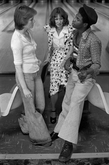 Black British boy chatting up a couple of teenage white girls, a group of teenagers hanging out in bowling alley Stevenage Hertfordshire 1970s They are wearing fashionable flared trousers known as Bell Bottoms. England 1975 UK HOMER SYKES
