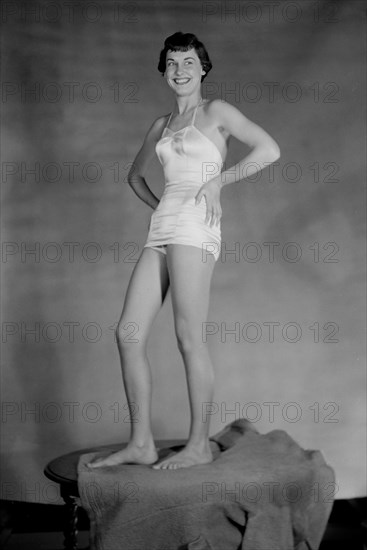 Young woman models a girdle while standing on a table, ca. 1955.