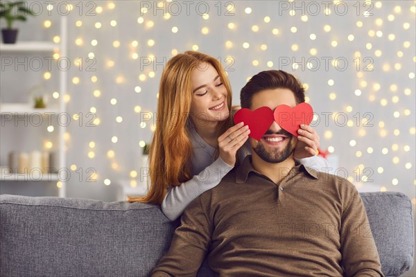 Woman covering boyfriend's eyes with red heart-shaped cards on Saint Valentine's Day