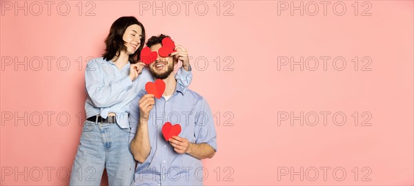 Flyer. Young and happy man and woman holding greeting cards shaped hearts isolated on pink trendy color background. Emotions, youth, love and