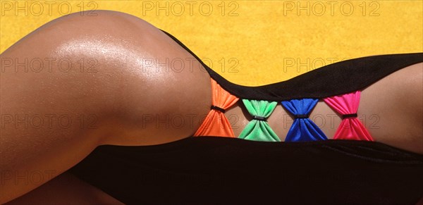 Historical 1988 archival model released abstract view of female  body curve shape created from a 1980s high cut on the thigh for woman fashion swimwear garment purchased in UK featuring close up detail of coloured bows connecting each black side of swimsuit against background of yellow towelling 80s archive outdoor image of how we were when photographed in Spanish holiday sunshine