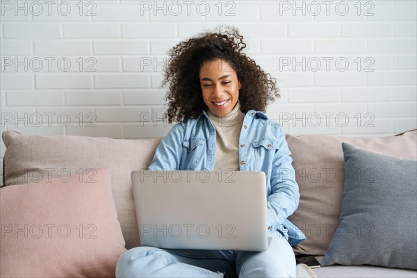 Happy african teen girl using laptop computer sitting on sofa.