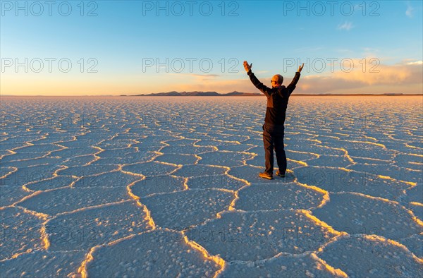 Sunset of the Uyuni Salt Flat Desert with Hexagon salt formations and a caucasian male tourist with stretched hands in the air, Bolivia.