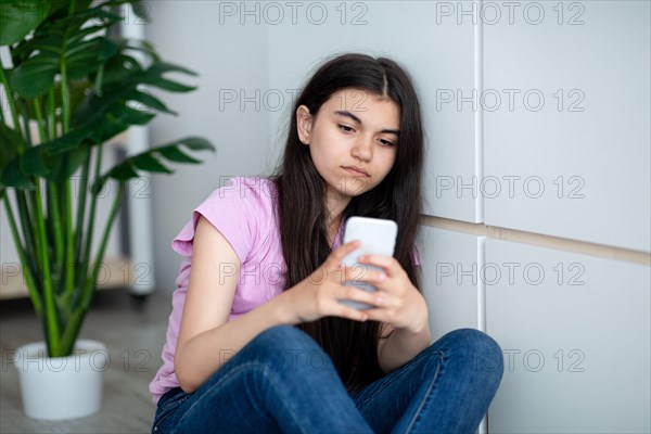 Depressed Indian teen girl with smartphone feeling lonely, looking at screen, sitting near wall at home