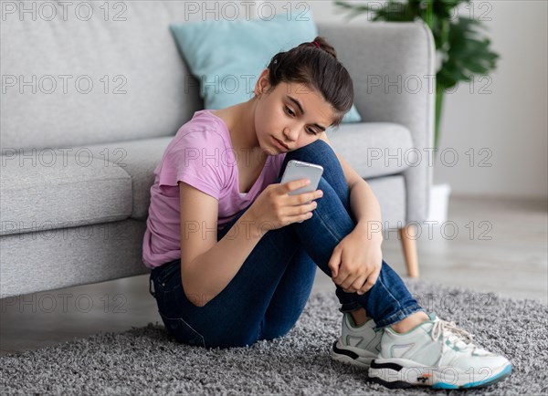 Unhappy Indian teen girl with mobile phone feeling sad, suffering from cyber bullying online, sitting on floor at home