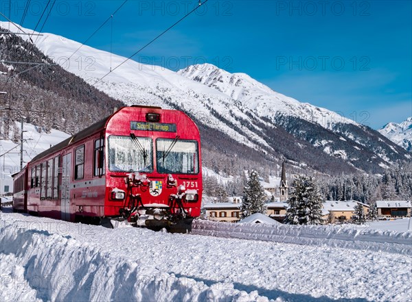 Cinuos-Chel, Switzerland - February 3, 2022: Red train riding between Chur and Tirano in Italy and crossing snow covered landscape at the village Chin