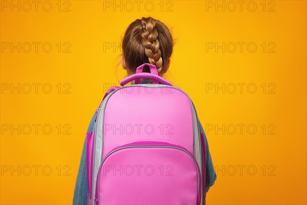 Little girl schoolgirl stands with her back on a yellow background. The child holds a satchel. School and education concept.