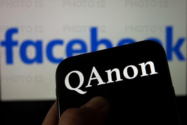 QAnon vs FACEBOOK. QAnon organisation logo seen on the smartphone which is placed on Facebook logos. Concept for ban of QAnon on social media.