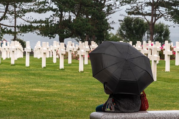 Colleville sur Mer, France - August 5, 2021: Woman with black umbrella meditating at the american Cemetery lined with crosses at Corlleville Sur Mer,