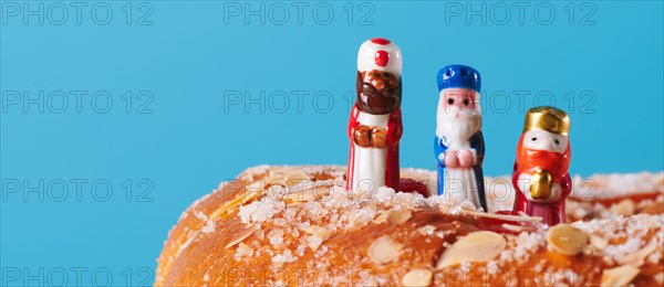 the three wise men, melchior, caspar and balthazar, on top of a roscon de reyes, the spanish king cake, on a blue background in a panoramic format to