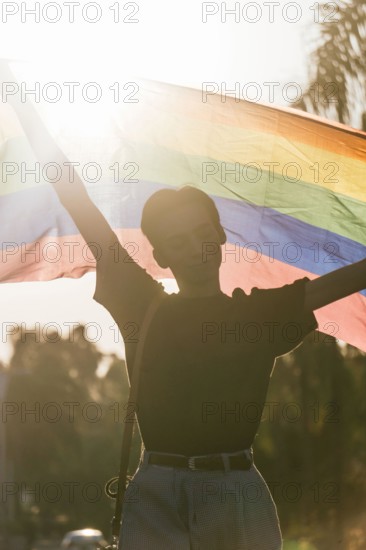 Delighted male gay standing with eyes closed raising rainbow LGBT flag during sunset in city