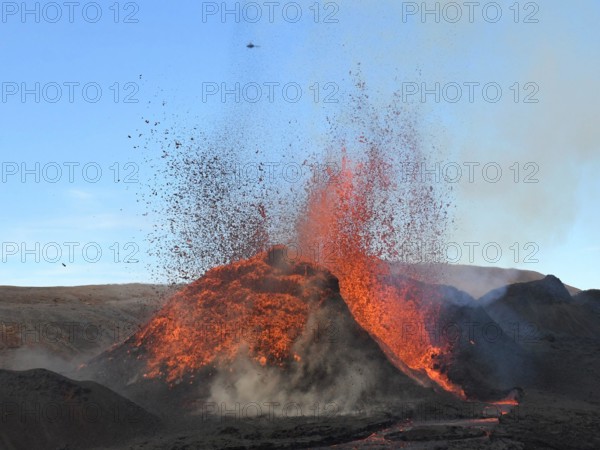 Volcanic vent at Fagradalsfjall, Iceland, erupting incandescent orange and red lava. Black lava in the foreground and blue sky in the background.