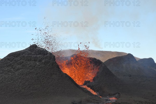 Volcanic vent at Fagradalsfjall, Iceland, erupting incandescent orange and red lava. Black cool lava in the foreground and blue sky in the background.