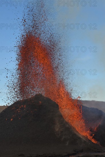 Volcanic vent at Fagradalsfjall, Iceland, erupting orange and red lava. Black cooled lava in the foreground and blue sky in the background.