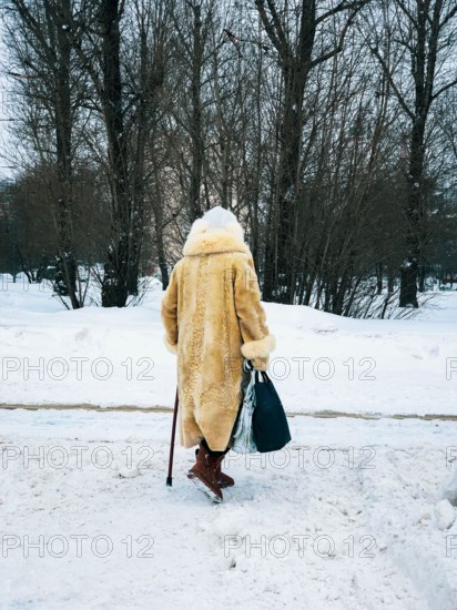 Winter. Back view of elderly women in fur coat with bags walking along snow with cane. Seniors, lifestyle concept