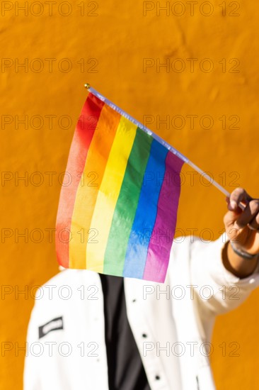 A young adult holds a multicolored rainbow flag in front of a bright yellow background