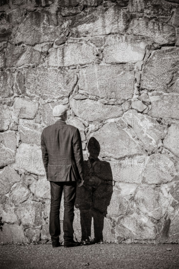 Old man standing in front of stone wall. Sunshine and heavy shadow. Building exterior with copy space.