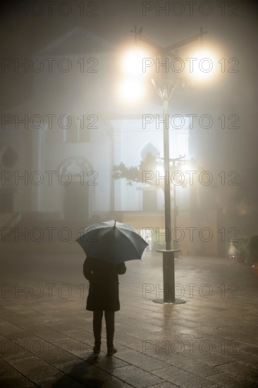 Person with umbrella standing under street lamp in front of the duomo in rain and fog at night, Ravello, The Amalfi Coast, Campania, Italy, Europe