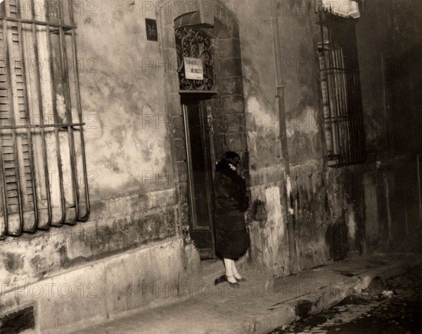 A woman stands by the door of a house in the port city of Marseille, France, in 1929, with a sign reading 'furnished rooms' above the door. Marseille, France, 1929.