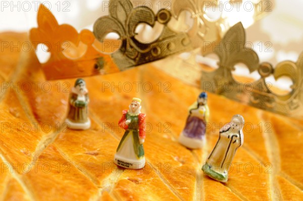 A golden, flaky galette des rois is on a yellow plate alongside traditional porcelain figurines and a paper crown. French Epiphanie