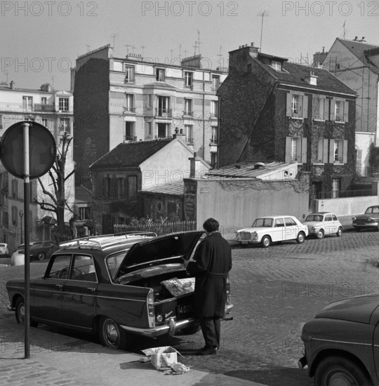 Pariser Bilder [The street life of Paris]  Man at opened trunk, with in the background Cabaret Au Lapin Agile on Rue des Saulfs in Montmartre Date: 1965 Location: France, Paris Keywords : cars, restaurants, nightclubs, street images