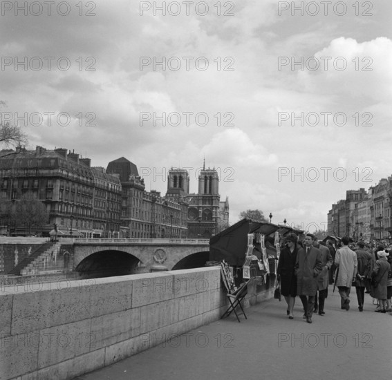 Pariser Bilder [The street life of Paris]  Passengers along the bookstalls at the Seine Annotation: The booksellers with a stall along the Seine are called bouquinistes. They sell old books and prints from large chests attached to the parapet Date: 1965 Location: France, Paris Keywords: markets, rivers, street statues, pedestrian Institution name: Notre Dame