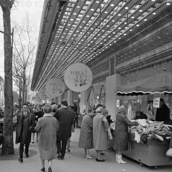 Pariser Bilder [The street life of Paris]  Shopping audience along the display of a clothing store Date: 1965 Location: France, Paris Keywords: clothing, street sculptures, shops