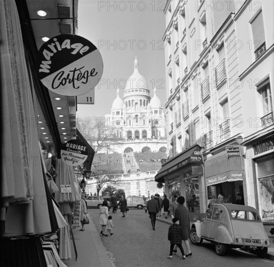 Pariser Bilder [The street life of Paris]  Shopping street with a view of the Basilique du Sacré-Coeur Annotation: The Basilique du Sacré-Coeur (1875-1914) was built on the occasion of the deaths that fell during the Franco-German War of 1870-1871, the design was by architect Paul Abadie Date: 1965 Location: France, Paris Keywords: basilicas, churches, street sculptures, shops Personal name: sacre coeur Institutional name: Sacré Coeur