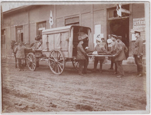 Wounded soldier on a stretcher, being loaded by stretcher bearers into a horse drawn ambulance. They are parked outside a building with Red Cross flags, the road is a wide mud track.