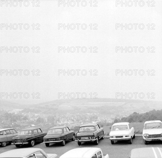 A number of cars are parked in a parking lot on a hill. Down in the valley is a community. Gandreas Uddling commented on the picture via the Digital Museum February 15, 2013: "View from France!" Car parking France 1960s.