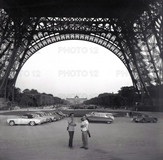 1961, historical, cars of the era parked underneath the Eiffel Tower in Paris, France, with two men standing in the open area under the giant metal structure, built for the 1889 Paris Exposition. Originally intended to be temporary structure, the wrought iron tower was due to be demolished in 1909, but was saved from destruction, due to its use as a radiotelegraph station. It is now one of the world's most iconic landmarks.