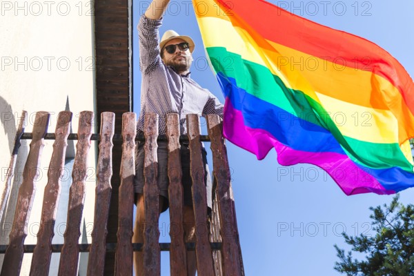 Man in hat holding lgbt rainbow flag in his hands while standing on the balcony.
