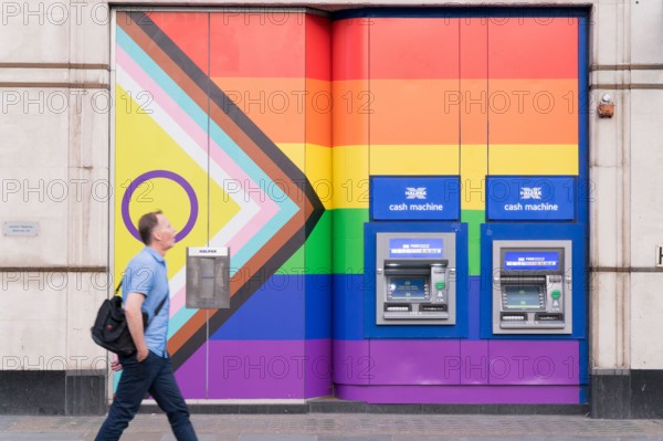A man walks past two cash machines on the wall painted with rainbow colours during celebration of Pride Month England UK