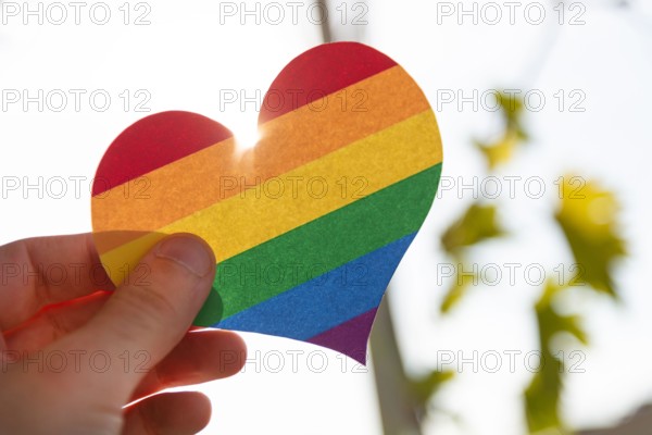 Hand holding a paper heart in LGBT rainbow colors with the sun rays shining around it, concept of LGBT, close-up